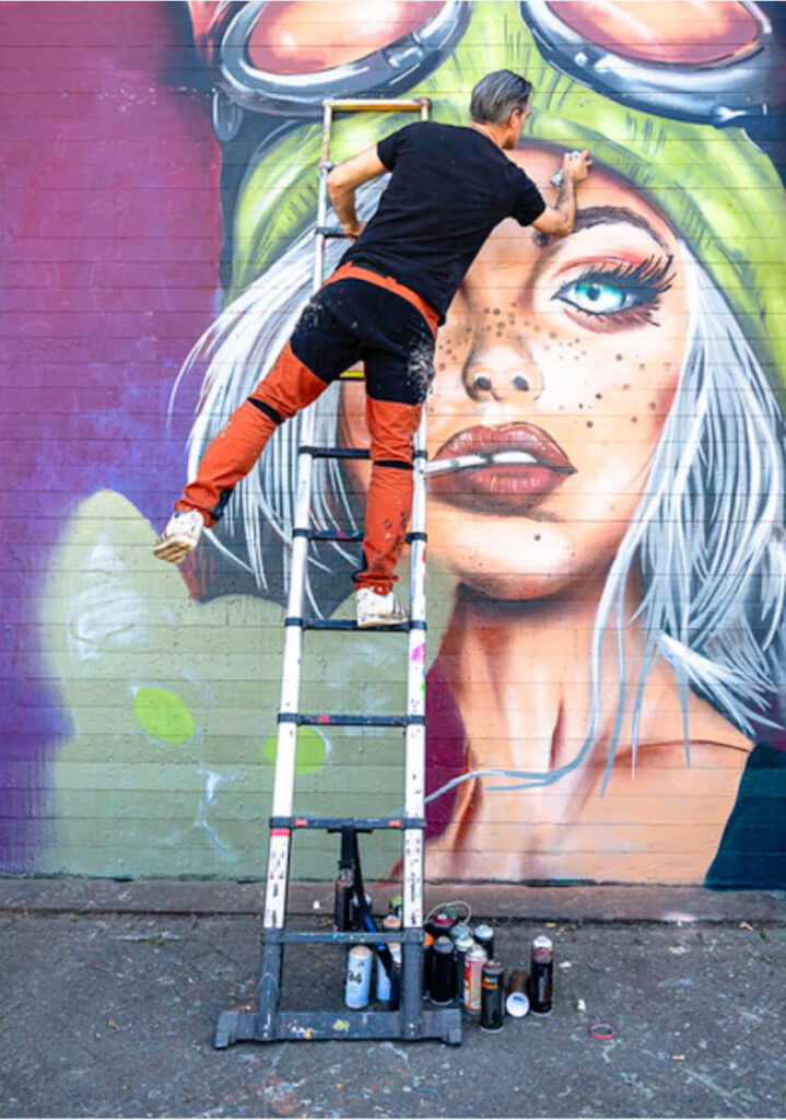 Theron on a ladder painting a vibrant mural of a woman with white hair and goggles on a brick wall, surrounded by spray paint cans.